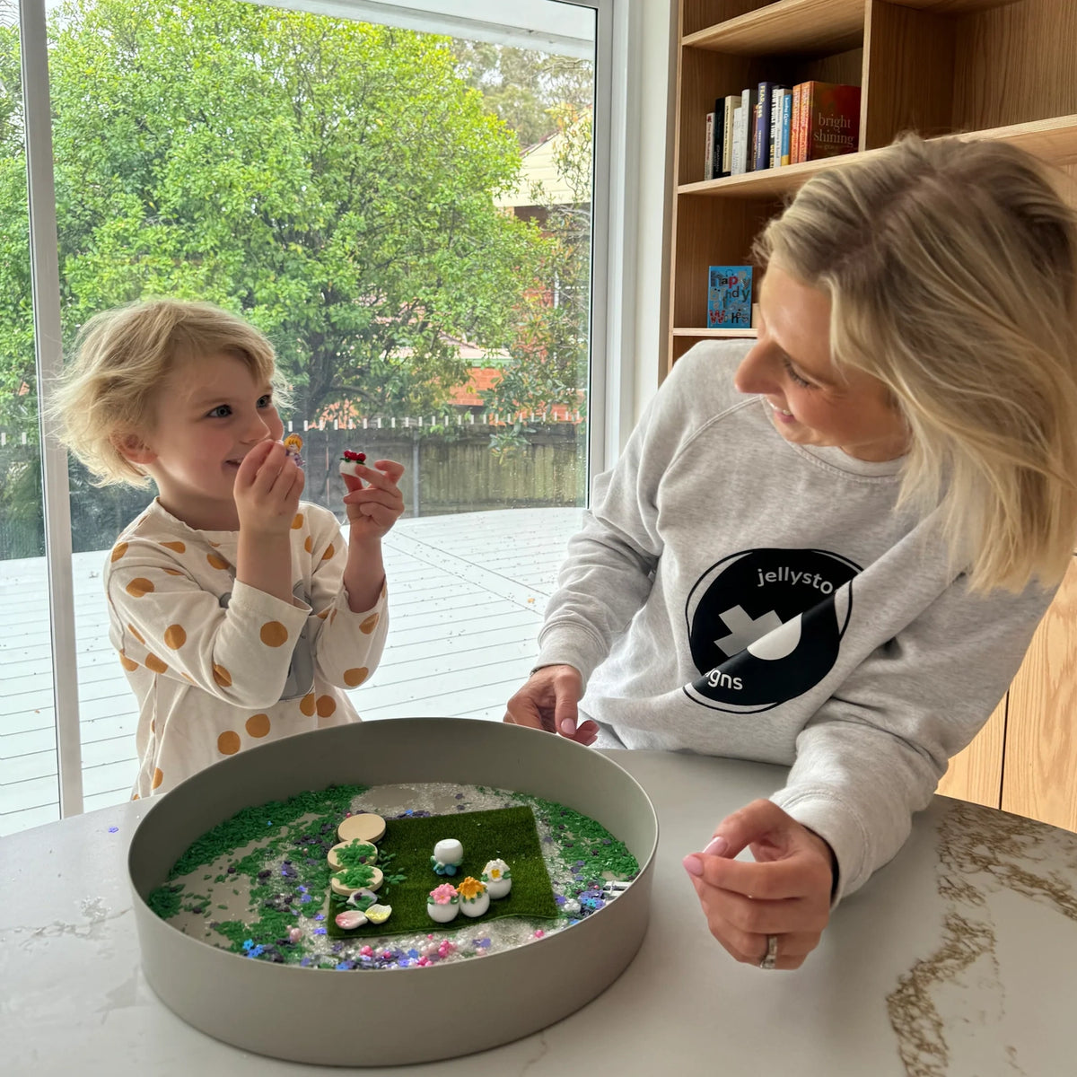 Woman and child playing together with a Jellystone Designs fairy garden sensory kit near a large window.
