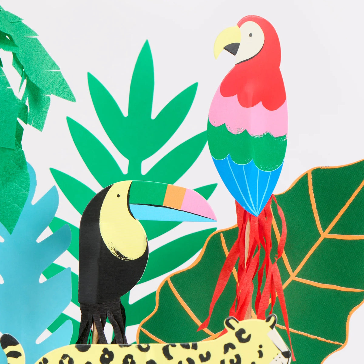Close-up of vibrant jungle-themed paper cake toppers featuring a toucan, parrot, cheetah, and tropical leaves.