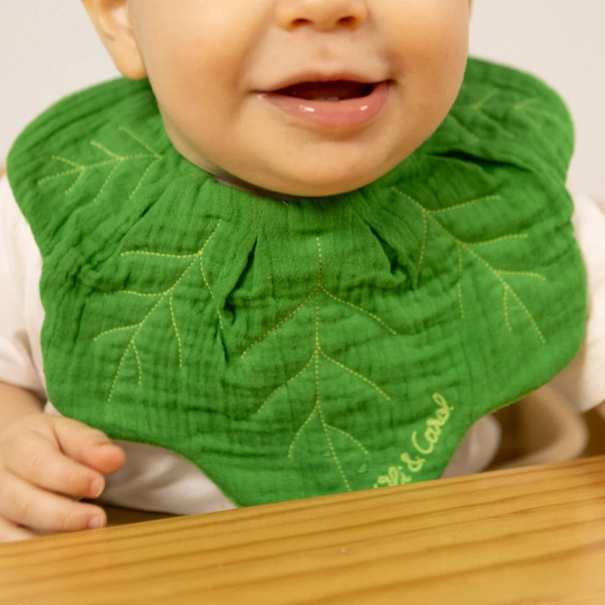 Smiling baby wearing a green leaf-shaped muslin bib with embroidered veins sitting at a wooden table.