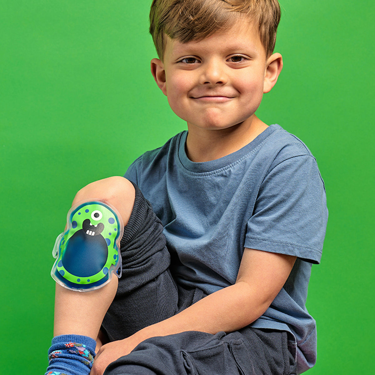 A smiling young boy sits against a bright green background, wearing a blue shirt. His knee is wrapped in a colorful, monster-themed ice pack.