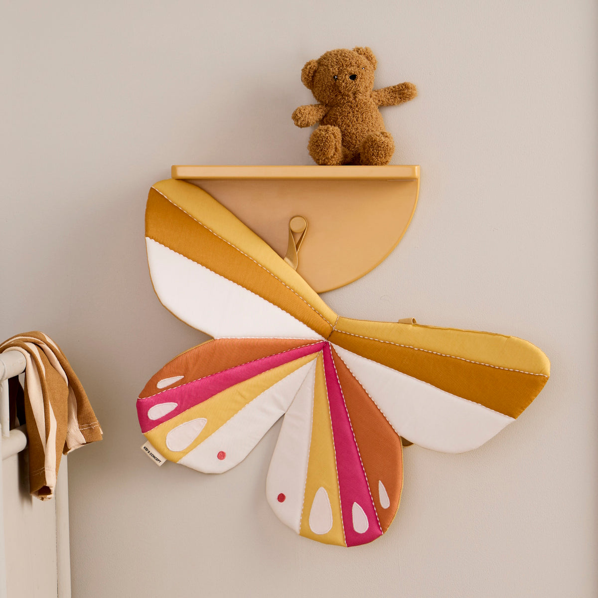 Fabric butterfly wings in mustard yellow, pink, and white hanging on a wall shelf with a teddy bear.