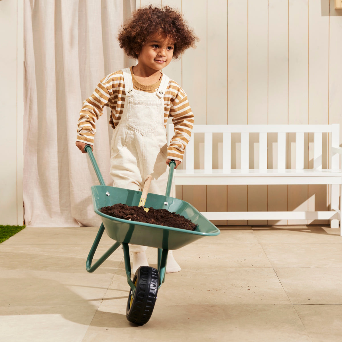 Young child pushing a green wheelbarrow filled with soil directly towards the camera.