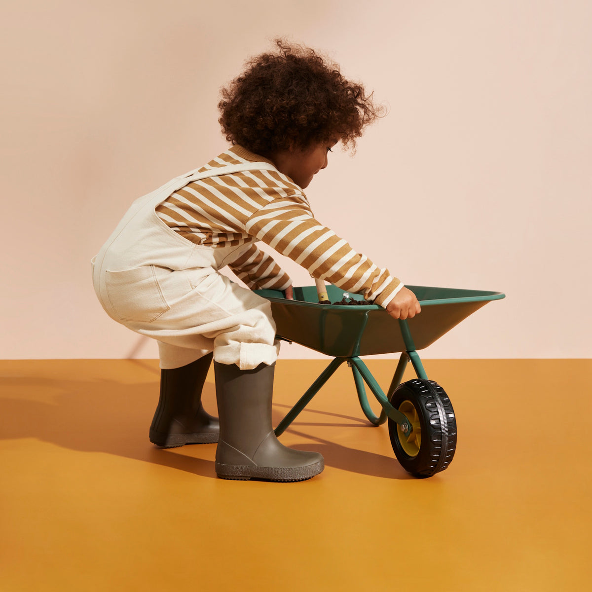Child wearing overalls and boots crouching beside a green wheelbarrow filled with soil.