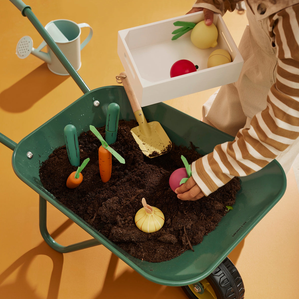 Child harvesting wooden play vegetables planted in the soil of a green wheelbarrow into a small white crate.