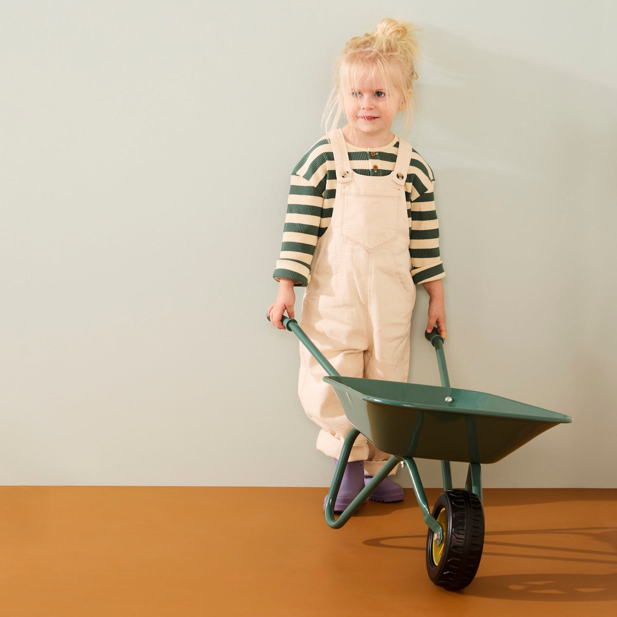 Young blonde child wearing cream overalls and a green striped shirt holding the handles of a green wheelbarrow.