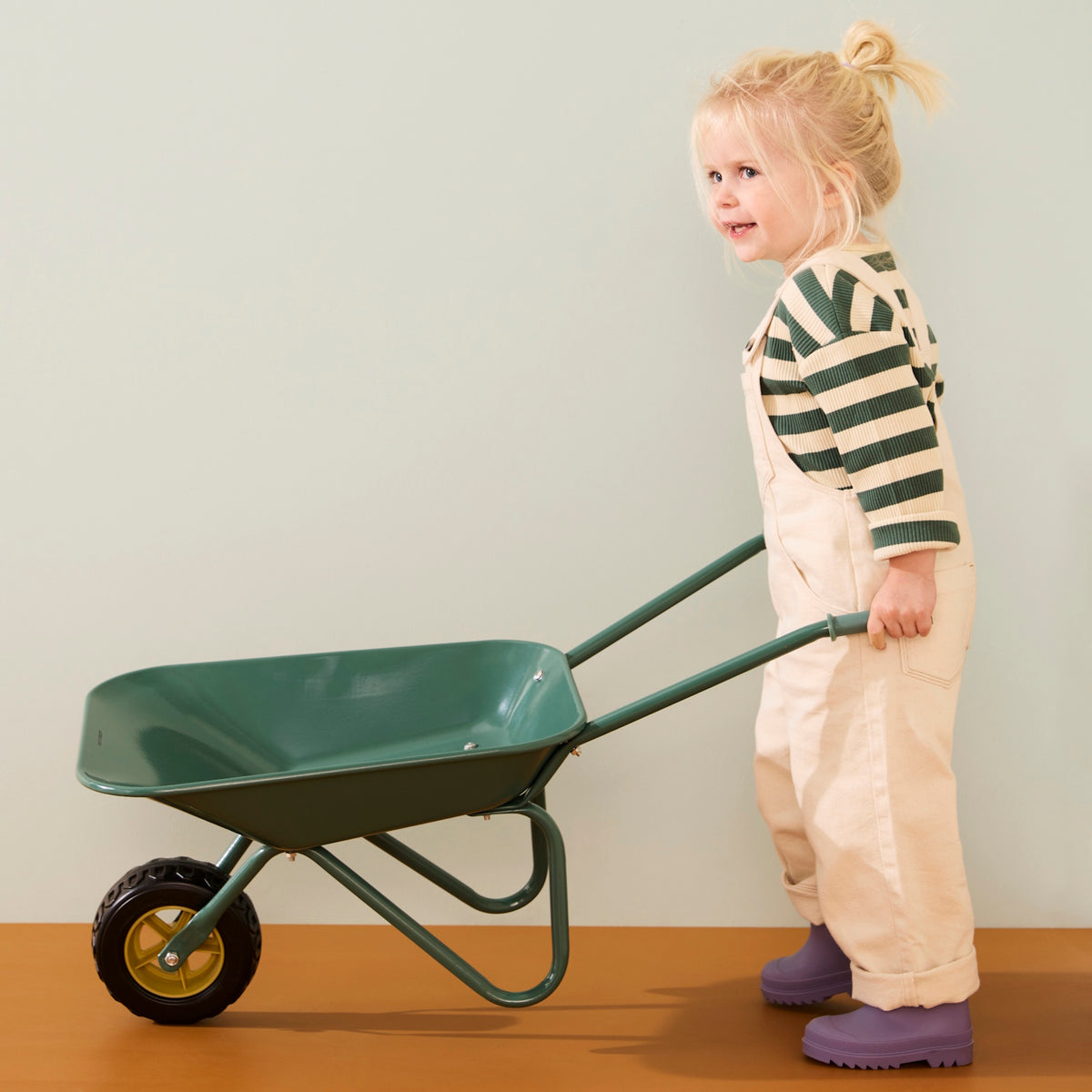 Young blonde child wearing cream overalls, a green striped shirt, and purple boots pushing a green wheelbarrow.