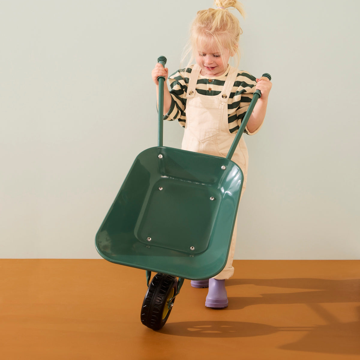 Young blonde child wearing cream overalls and purple boots holding up the handles of a green wheelbarrow.