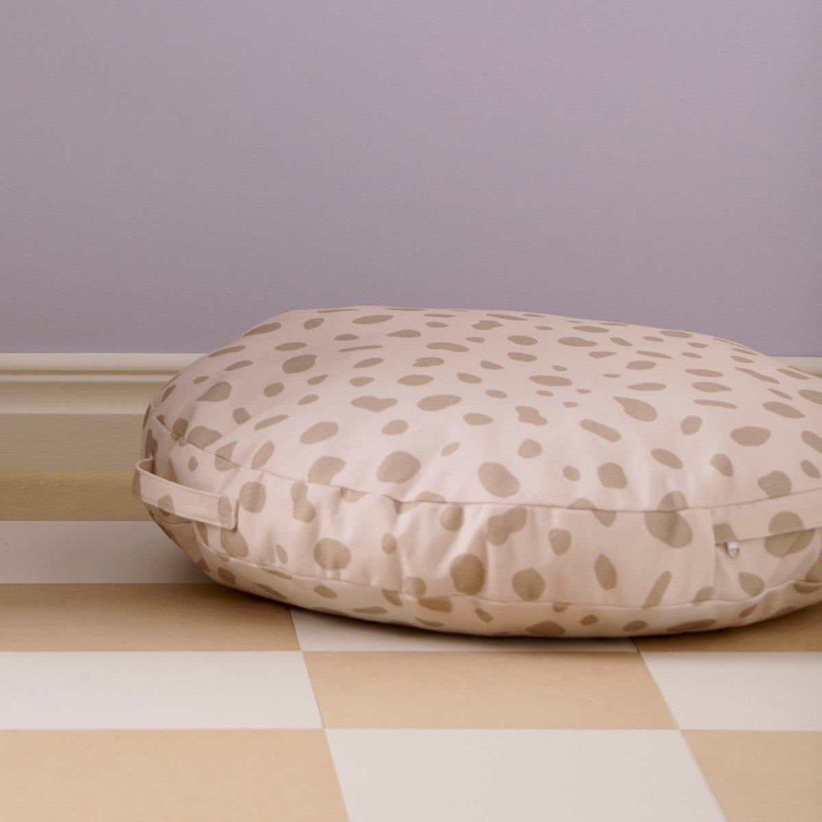 A round, beige polka-dot floor cushion resting on tiled flooring against a plain wall.