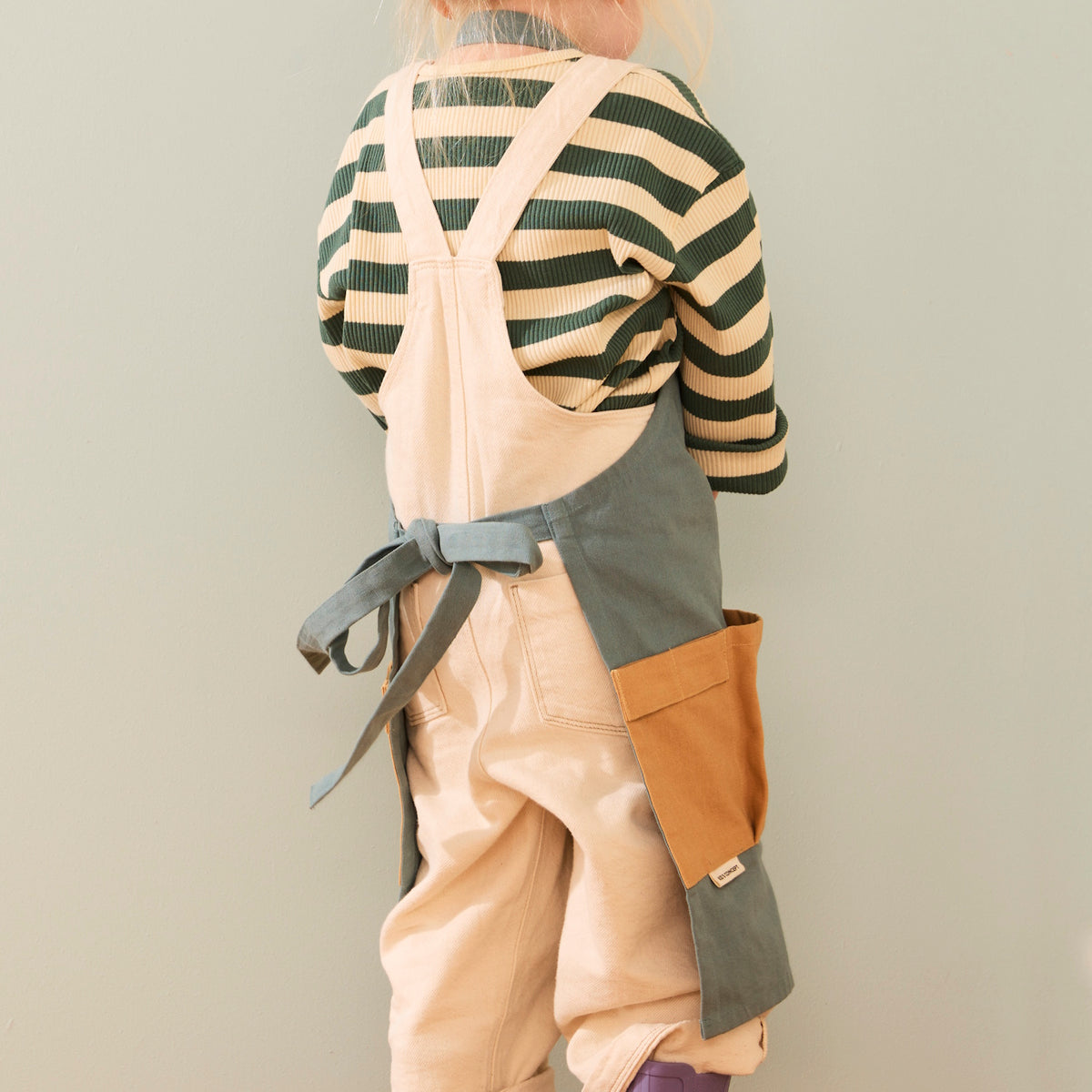 Back view of a child wearing a green apron tied around the waist over beige overalls.