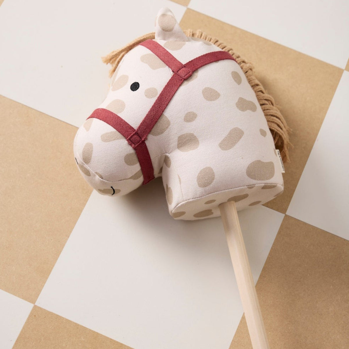 Plush hobby horse with beige spots and a red bridle, resting on a checkered floor.