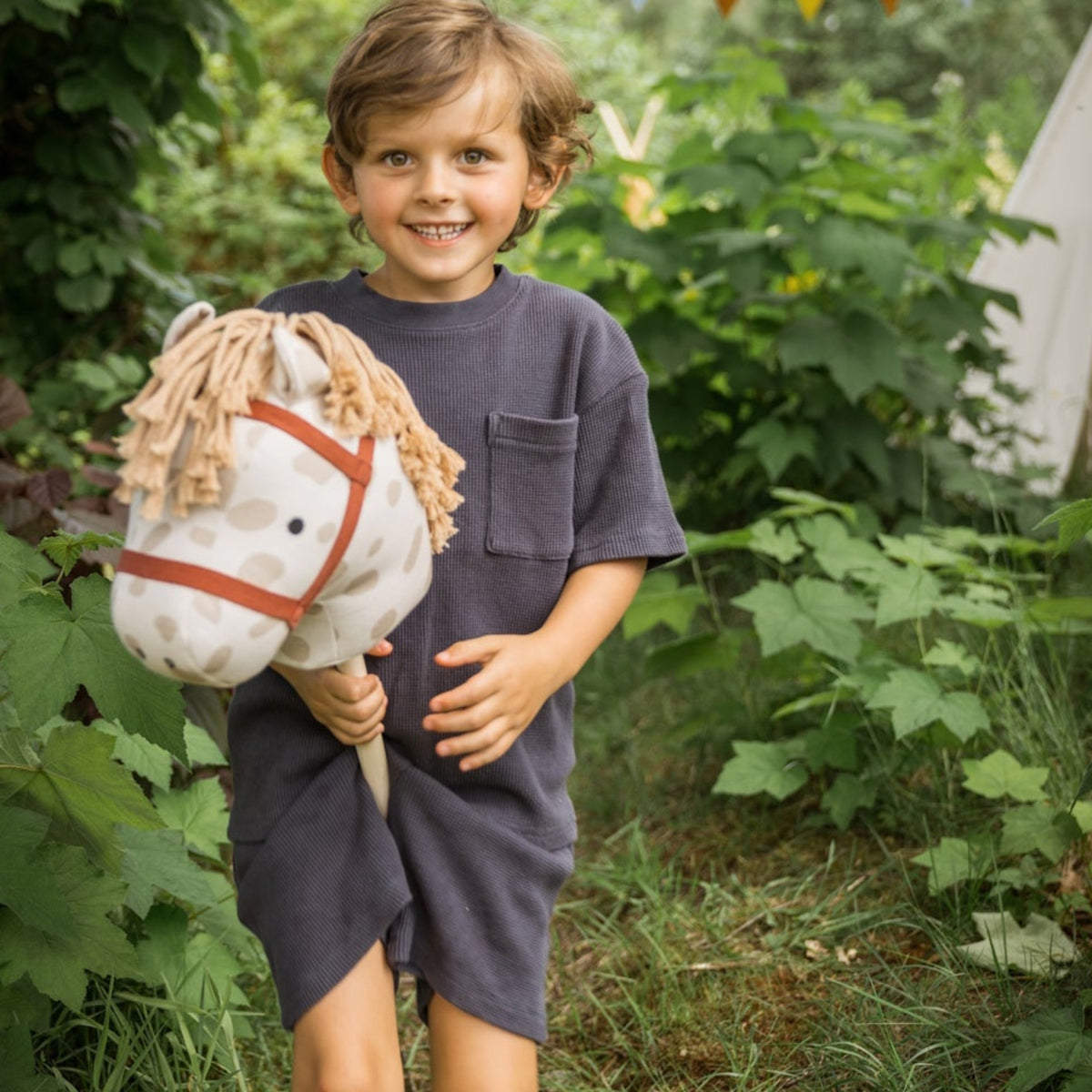 Smiling child holding a spotted stick horse in an outdoor garden with colorful party bunting.