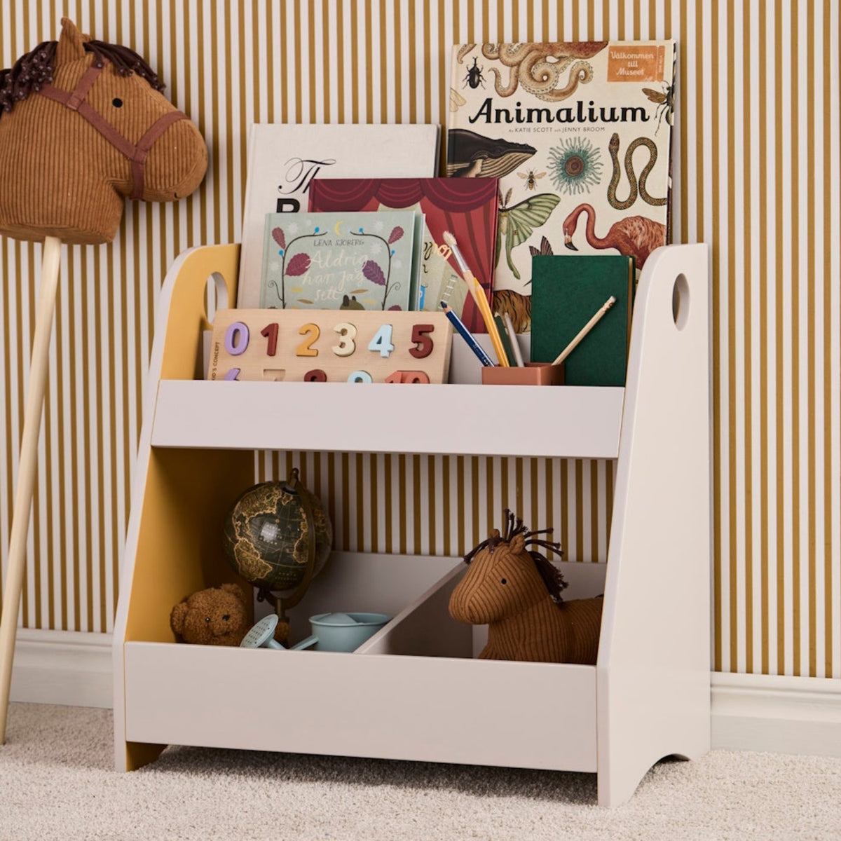Brown corduroy hobby horse next to a bookshelf filled with books and toys against striped wallpaper.