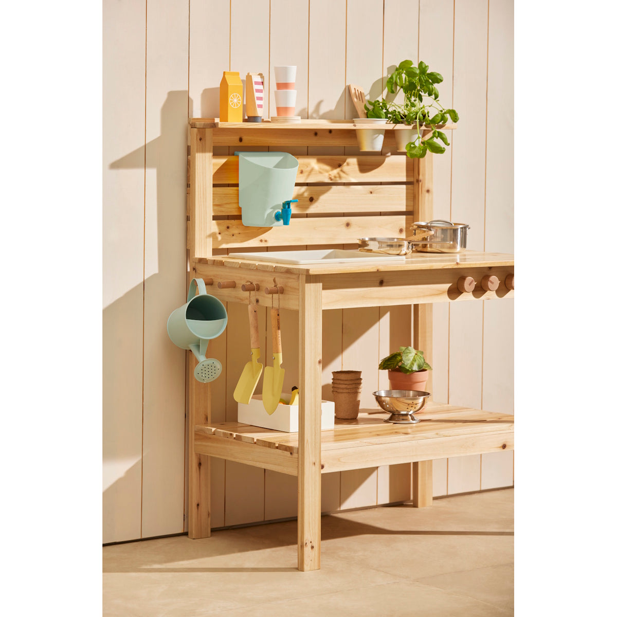 Wooden play mud kitchen equipped with play accessories, garden tools, and plants.