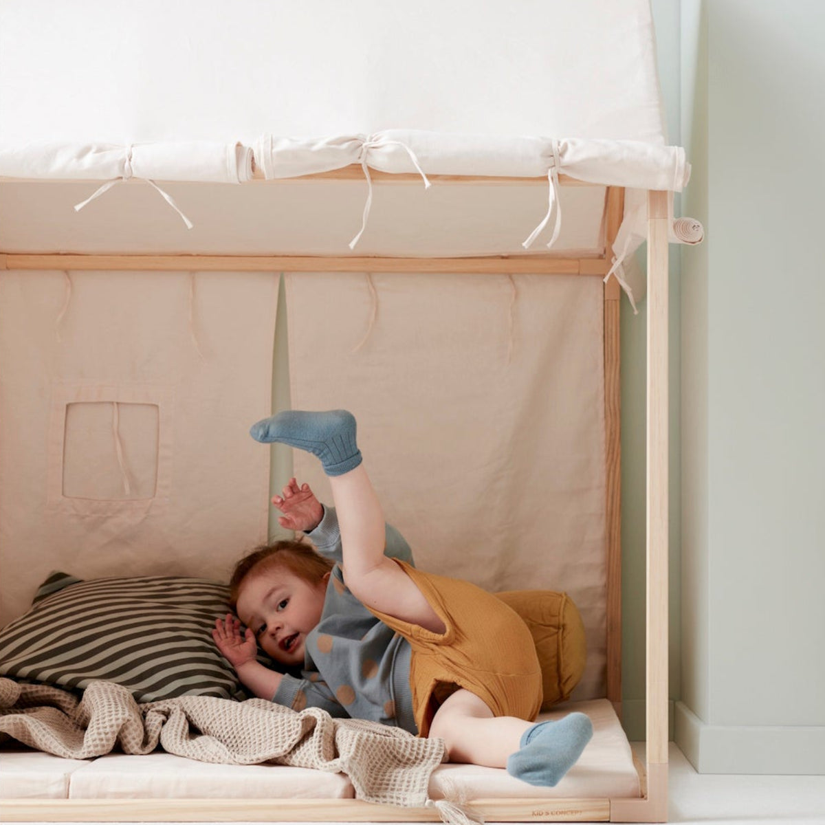 Toddler kicking feet up while lying inside an off-white play tent with cushions and a blanket.
