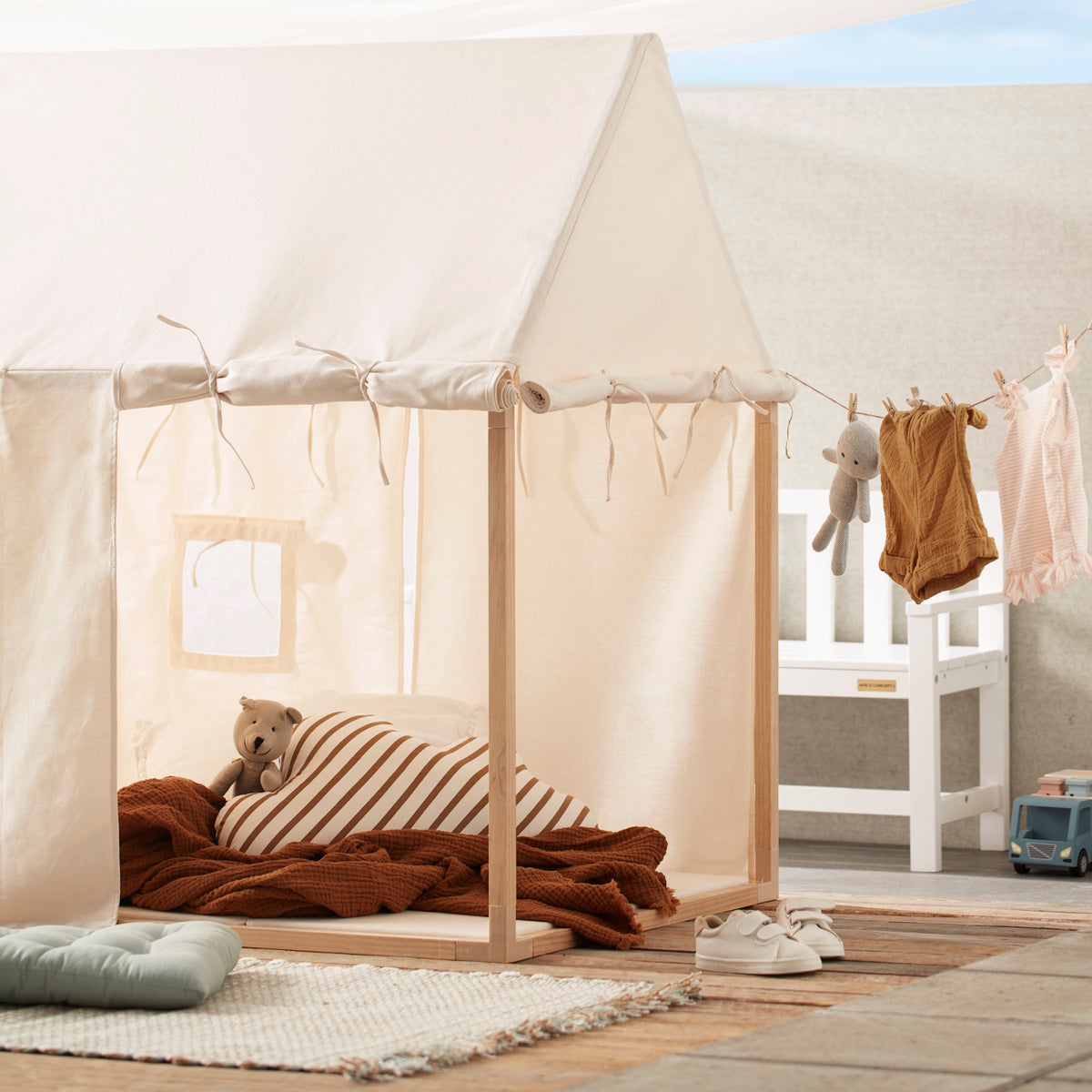 Off-white play house tent set up outdoors with cozy cushions and clothes hanging on a line nearby.