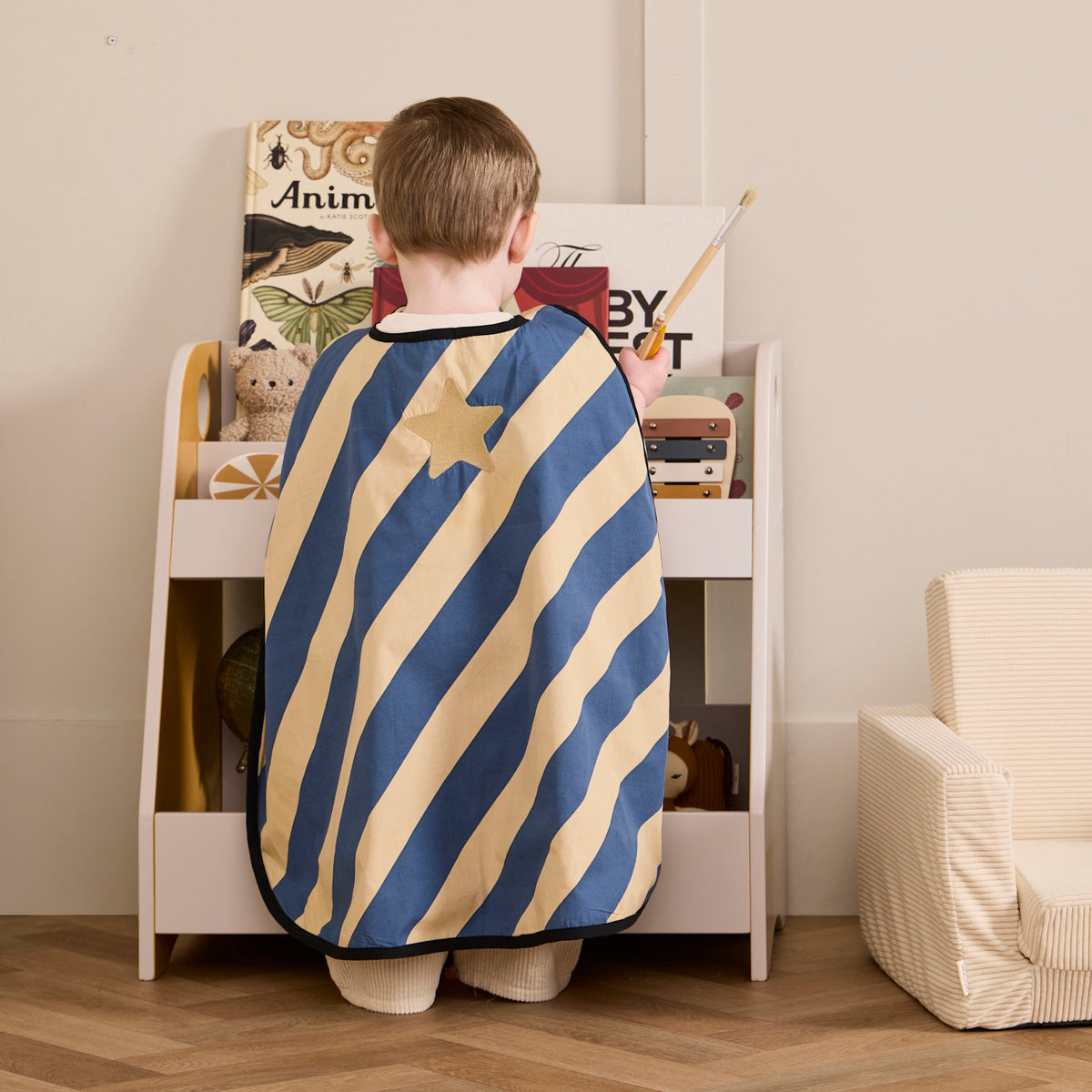 Young child from behind wearing a blue and yellow striped dress-up cape with a gold star badge.