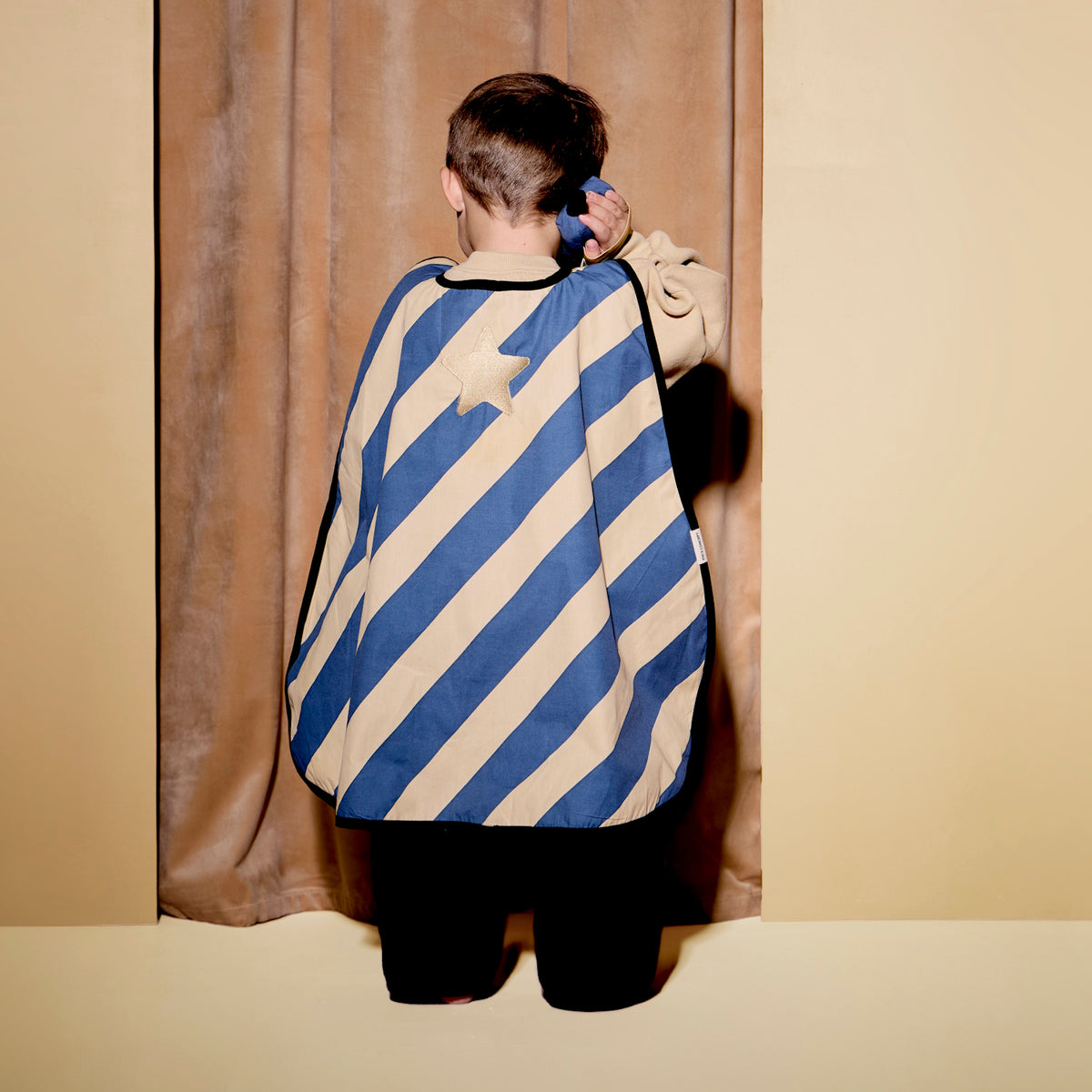 Child seen from behind wearing a striped dress-up cape, holding a toy walkie-talkie to their ear.