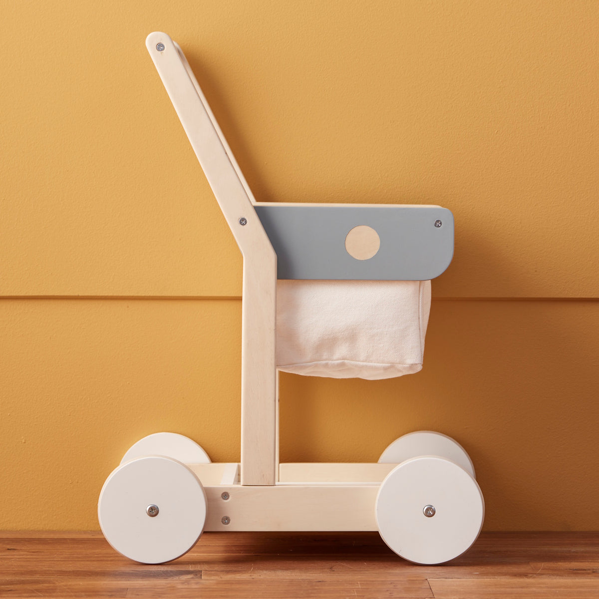 Side view of a wooden toy shopping trolley with a fabric basket against a warm yellow wall.