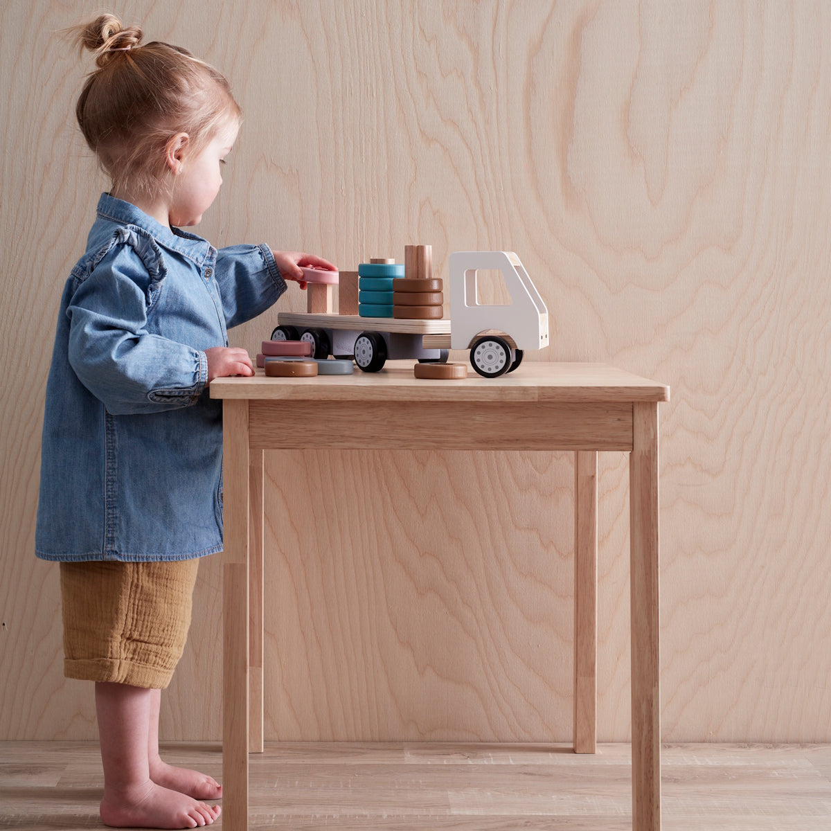 Toddler standing at a wooden table, playing with a wooden toy truck and stacking rings.