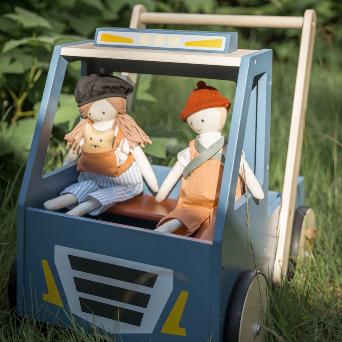Blue wooden toy walker truck carrying two fabric dolls outdoors on grass.