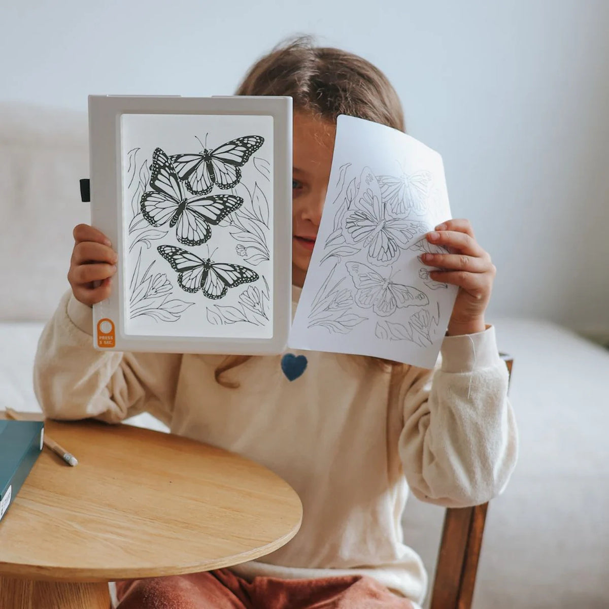 A child holding an illuminated drawing pad and a tracing paper showing butterfly drawings.
