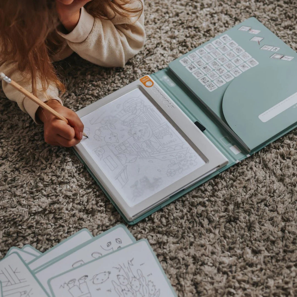 A child drawing with a pencil on an illuminated drawing pad while lying on a textured gray rug.