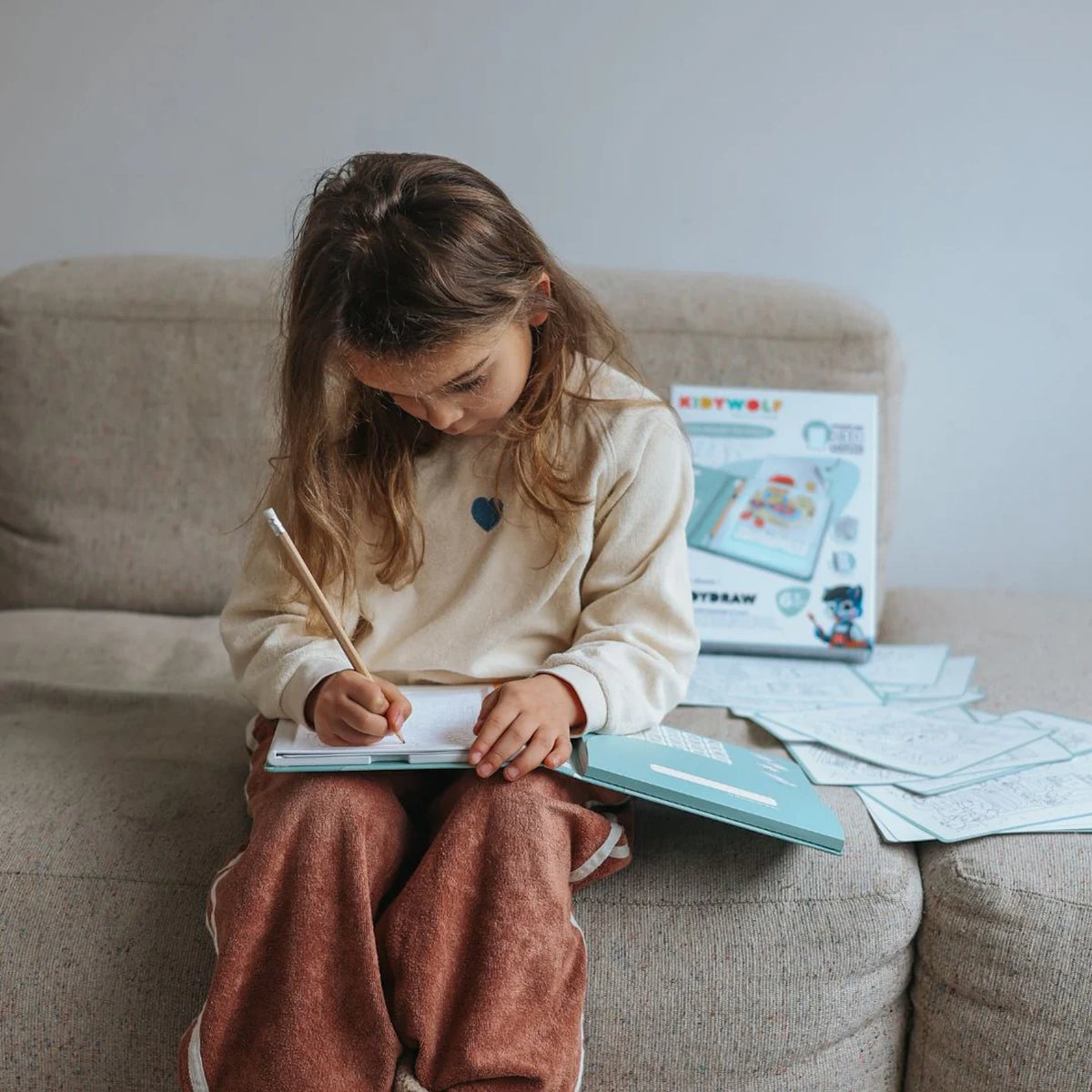 A young girl sitting on a couch, focused on drawing on an illuminated sketch pad with a pencil.