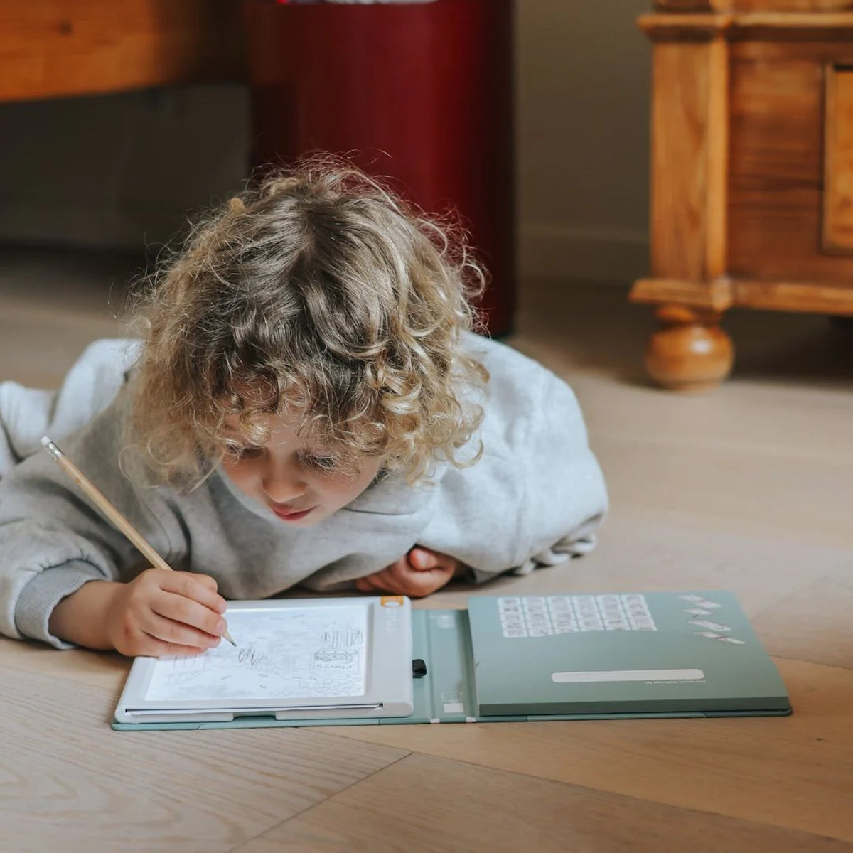 A young child with curly hair lies on the floor, drawing on a glowing light pad with a pencil.