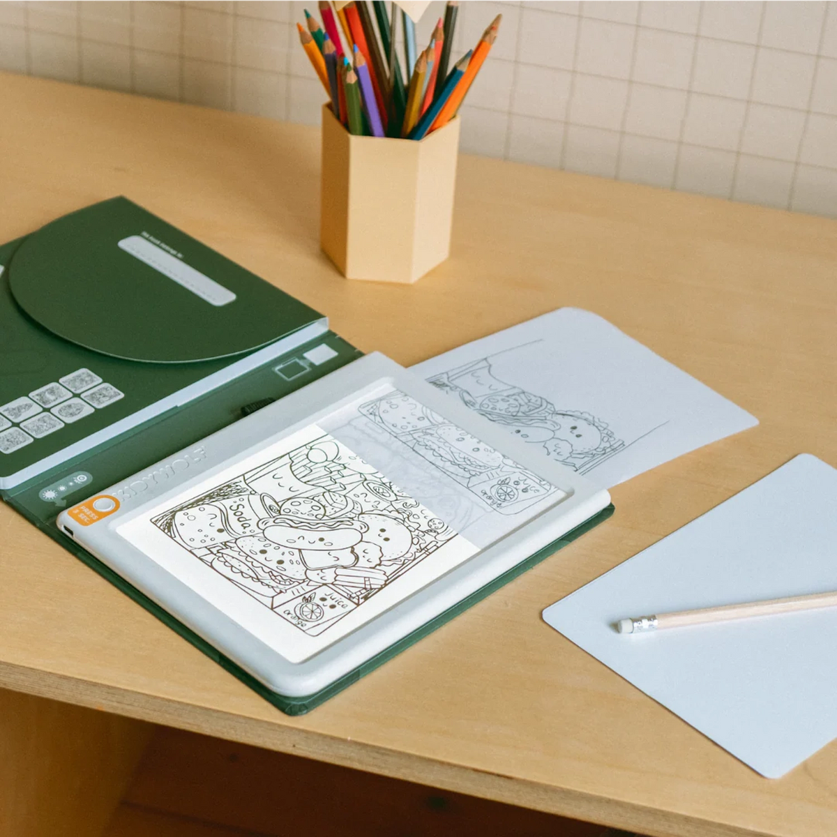 An illuminated light pad displaying a food drawing next to paper, a pencil, and colored pencils on a desk.