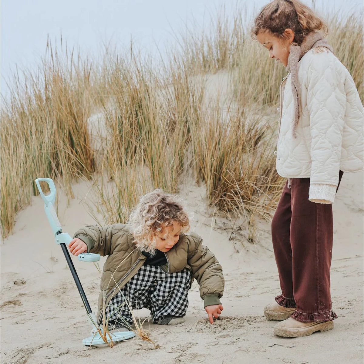 Two children playing with a toy metal detector on a sandy beach by tall grass.