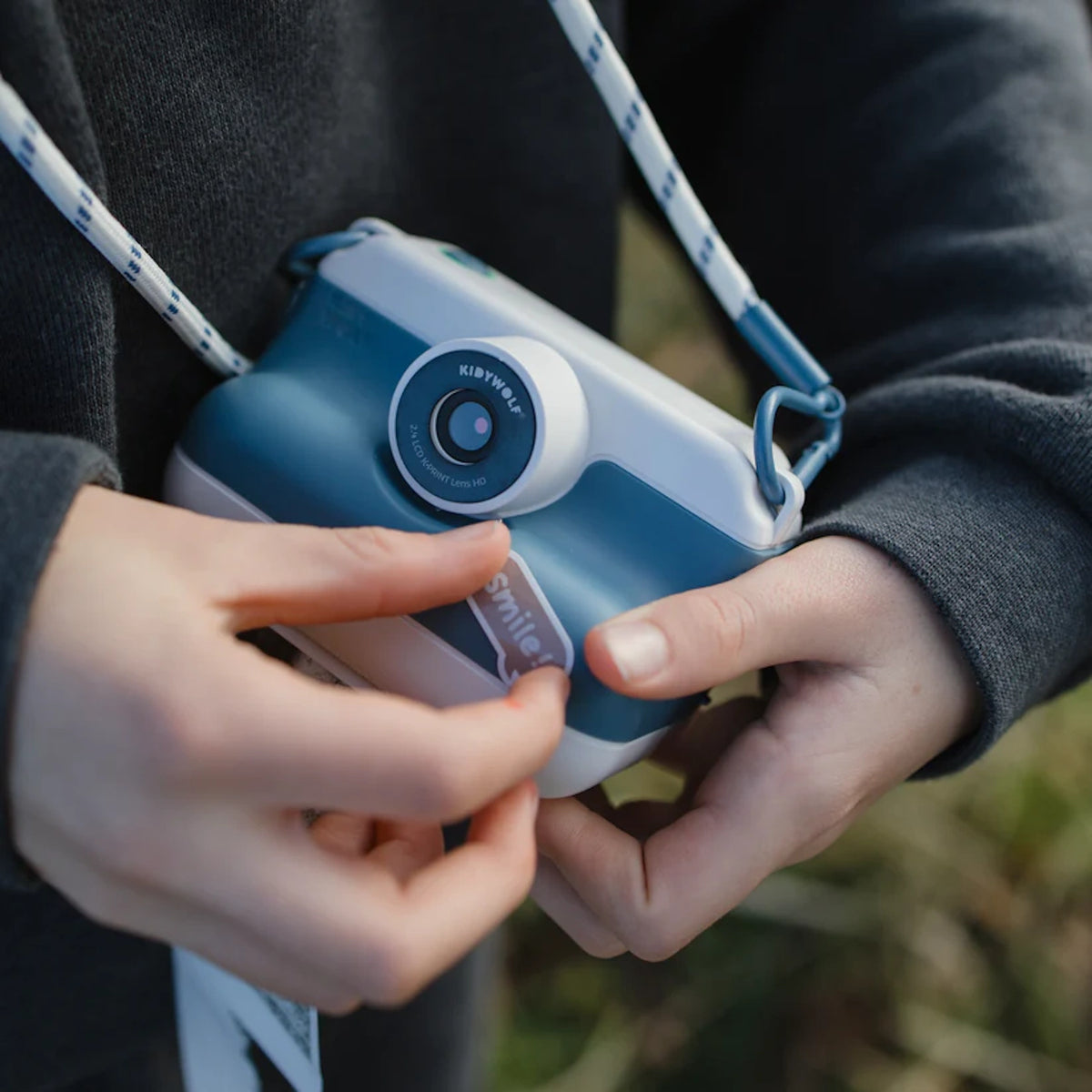 Child using a blue Kidywolf Kidyprint instant camera with thermal printer and "Smile" sticker decoration.