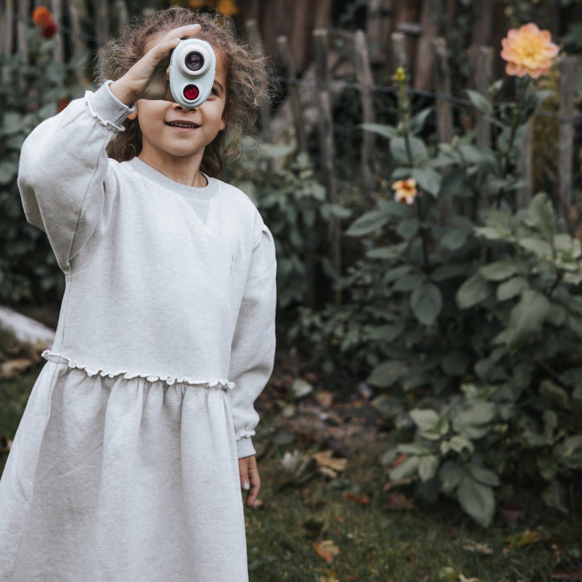 Young girl in a grey dress smiling while playfully looking through a toy camera in a garden, surrounded by green plants and orange flowers.