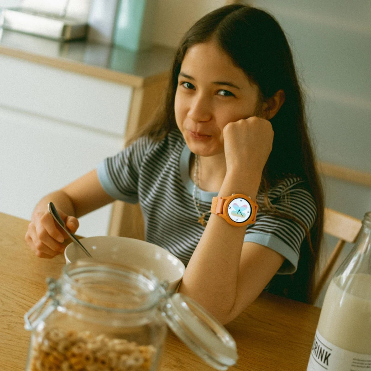 A young girl wearing a coral smartwatch eats breakfast at a wooden kitchen table.