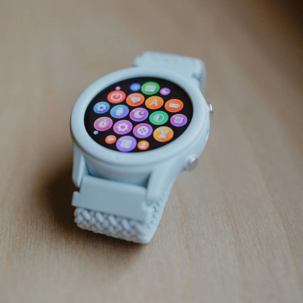 A light blue smartwatch with a round screen showing colorful app icons sits on a wooden surface.