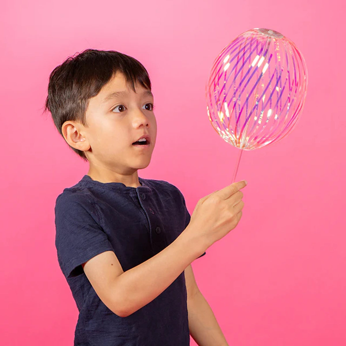A boy in a navy shirt looks amazed while holding a colorful, glowing balloon against a vibrant pink background. The scene conveys wonder and curiosity.