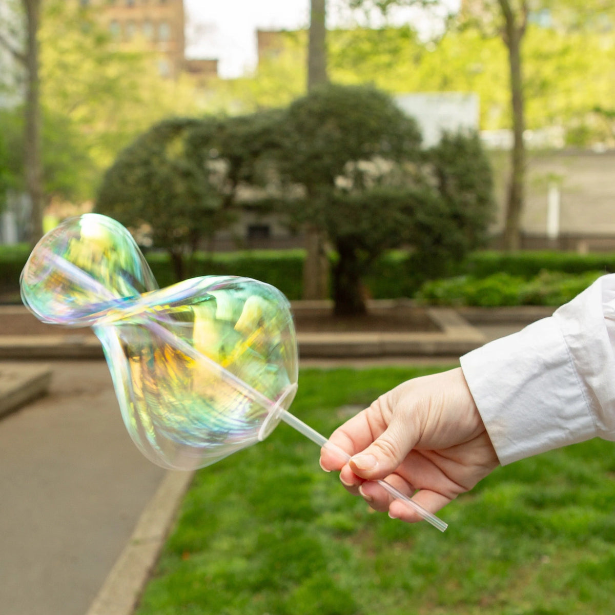 A hand in a white sleeve holds a straw with a large, iridescent soap bubble outdoors. The background shows greenery and blurred trees, creating a playful, light-hearted atmosphere.