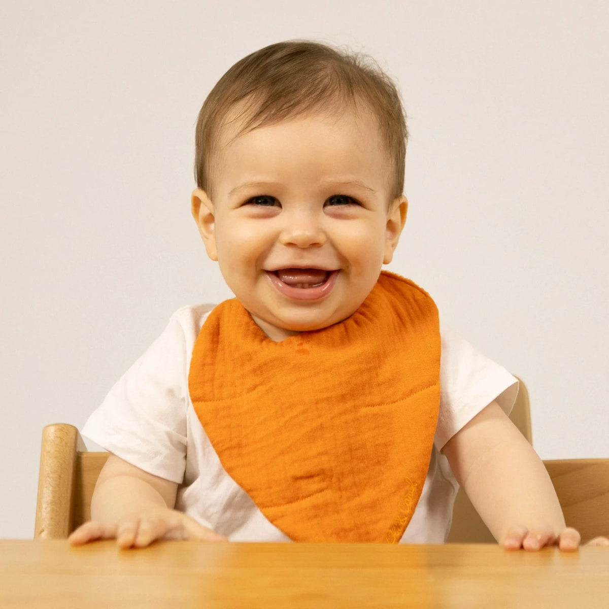 Smiling baby wearing an orange muslin bandana bib and white t-shirt, sitting at a wooden table against a plain background.