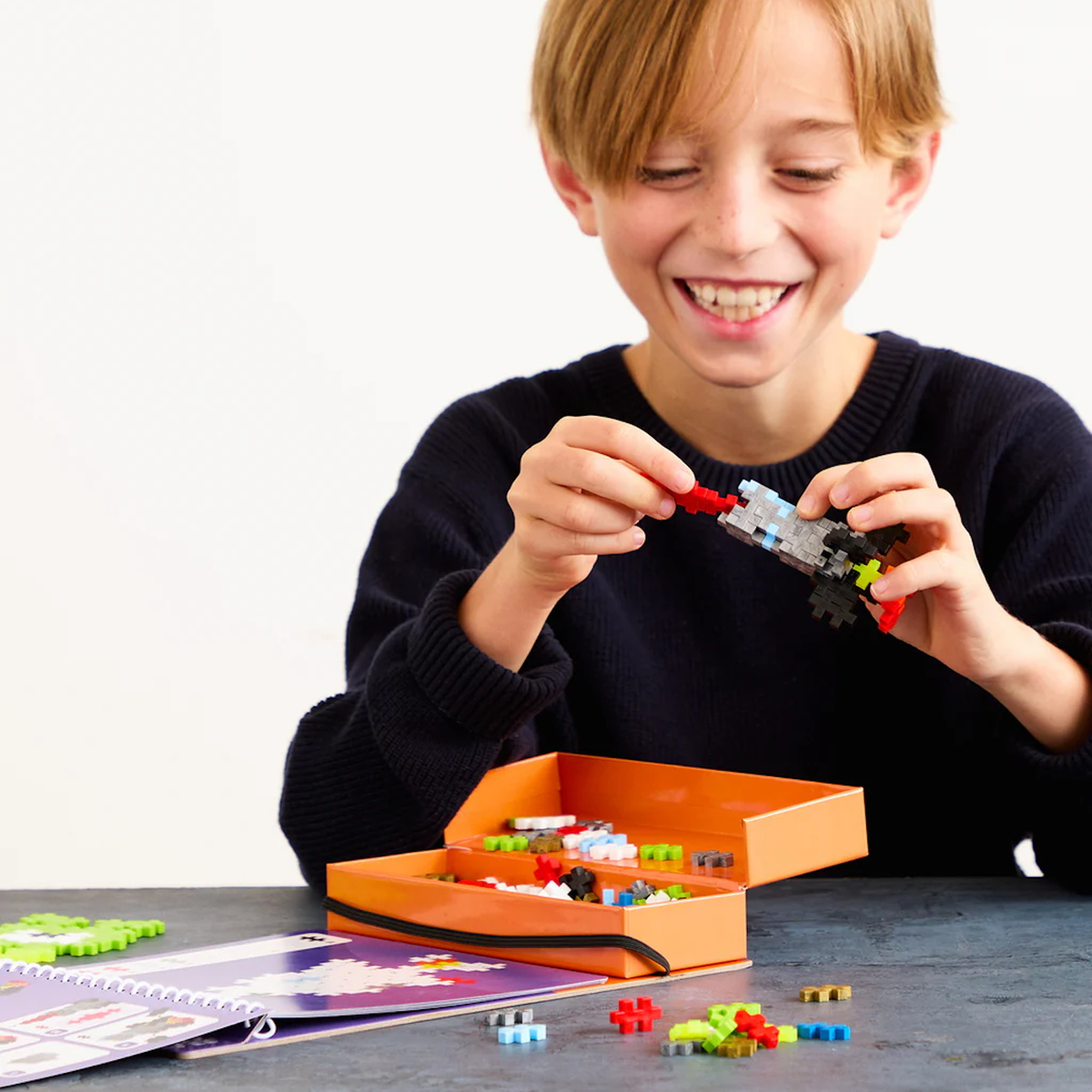Smiling child building a toy rocket with Plus-Plus blocks from a space-themed activity set on a blue table.