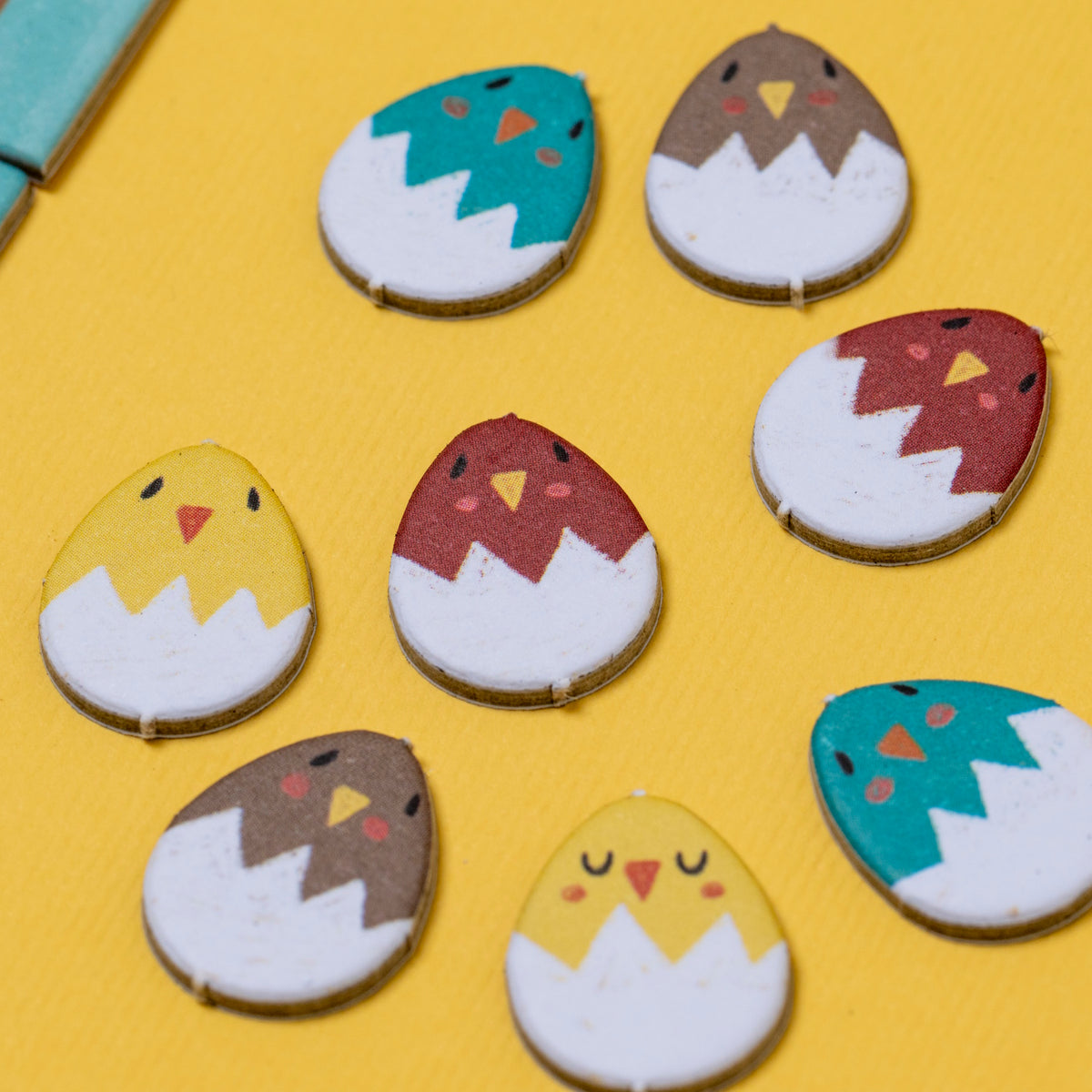 Colorful wooden Piu Piu counting game pieces shaped like chicks hatching from white eggshells on a yellow background.