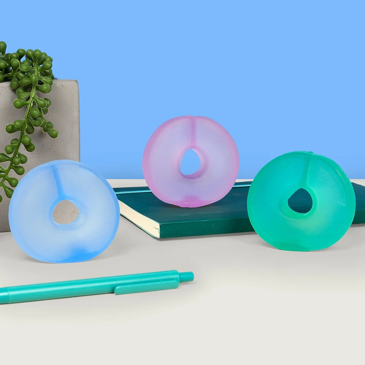 Three cool-toned gel stress loops displayed on a desk with a notebook, pen, and potted plant.