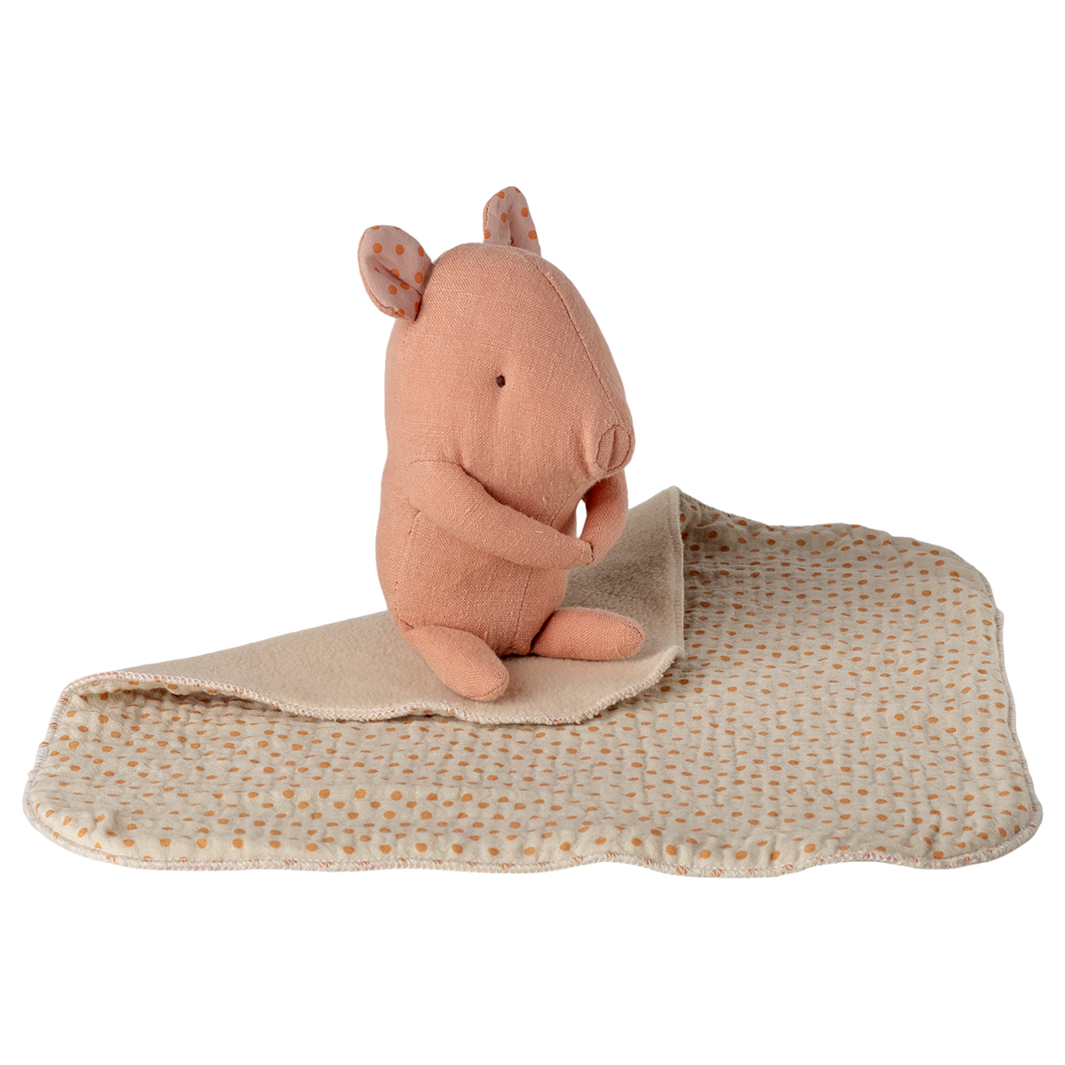 A soft pink stuffed animal resembling a small rodent sits on a beige blanket with brown polka dots, set against a white background. The tone is cozy.
