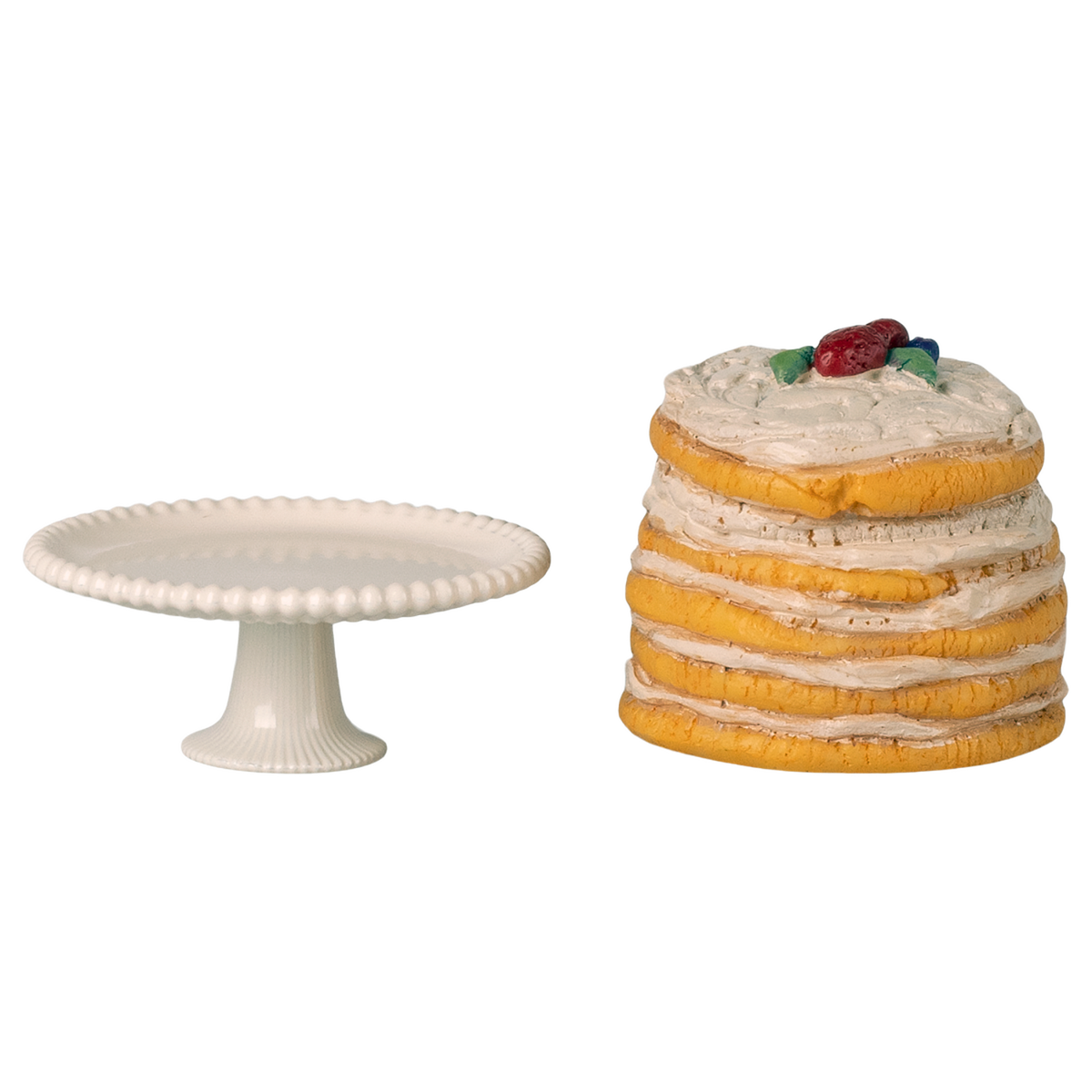 White cake stand next to a stack of pancakes topped with cream and berries, set against a white background. The scene feels whimsical and playful.