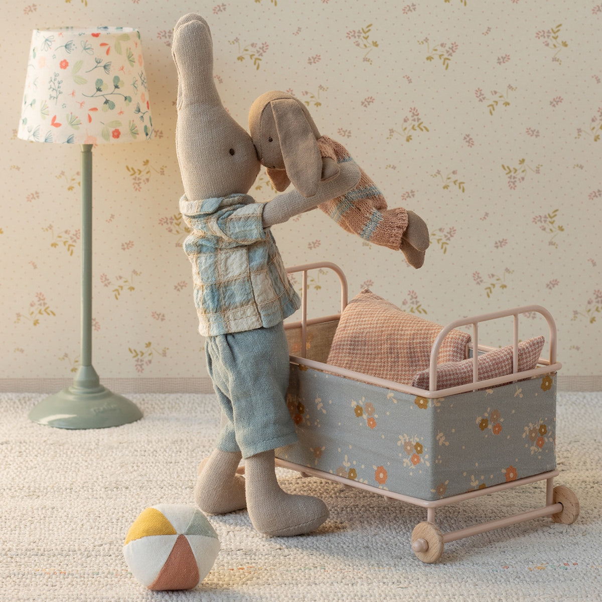 Maileg stuffed bunny in a blue plaid shirt lifting a small rabbit over a floral toy crib in a nursery setting.