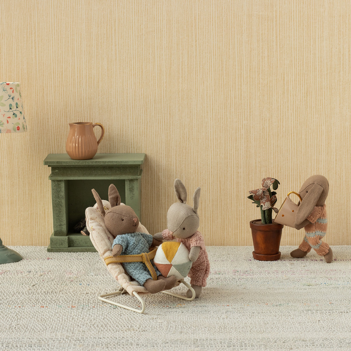 Three Maileg micro bunnies playing in a miniature room with a bouncer, ball, and watering can in a dollhouse setting.