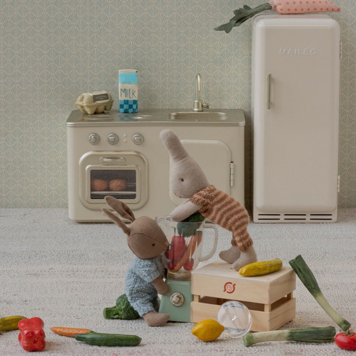 Maileg micro rabbits playing in a miniature kitchen with a blender, toy food, stove, and refrigerator.