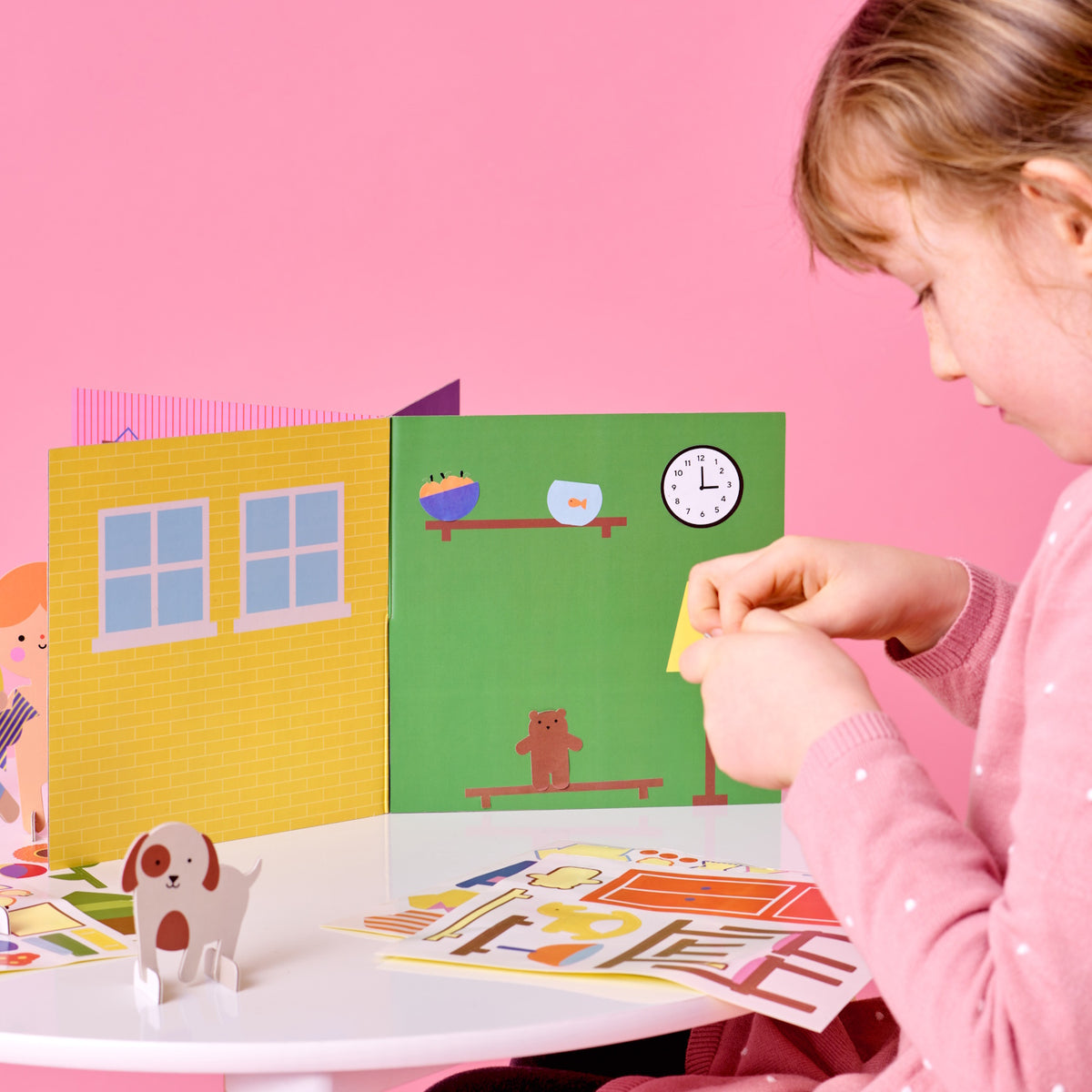 Young girl decorating a colorful 3D paper house playset with stickers on a white table.