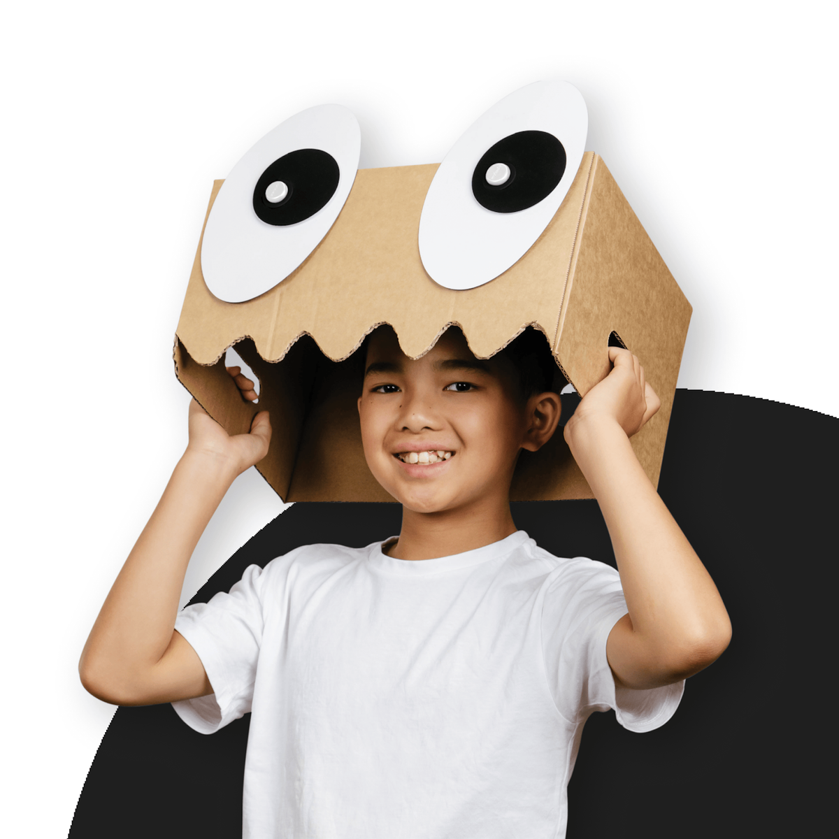Smiling boy wearing a cardboard box mask on his head with large googly eyes and a jagged cut mouth.