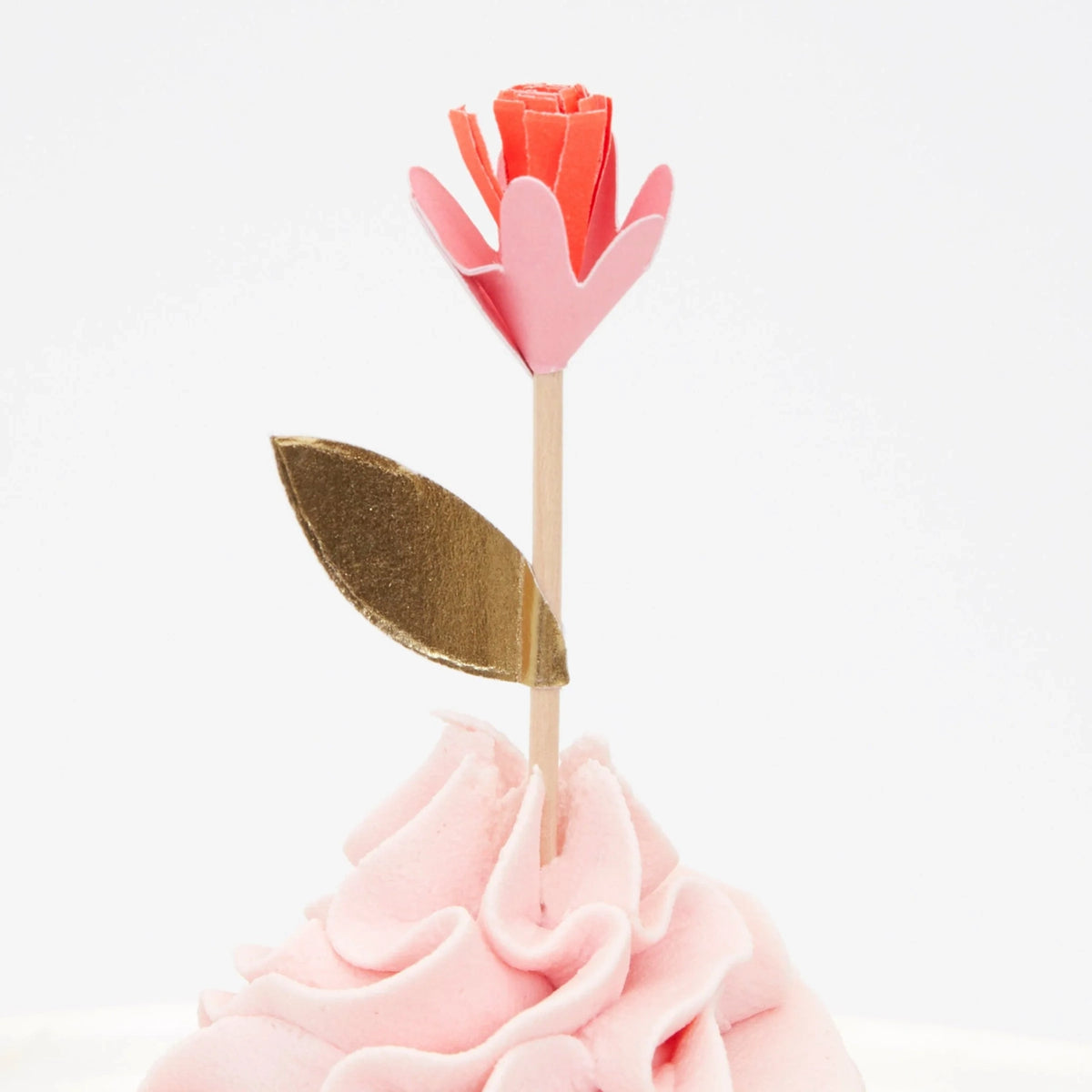 A paper flower cupcake topper with pink and orange petals and a gold leaf on a wooden stick, in pink frosting.
