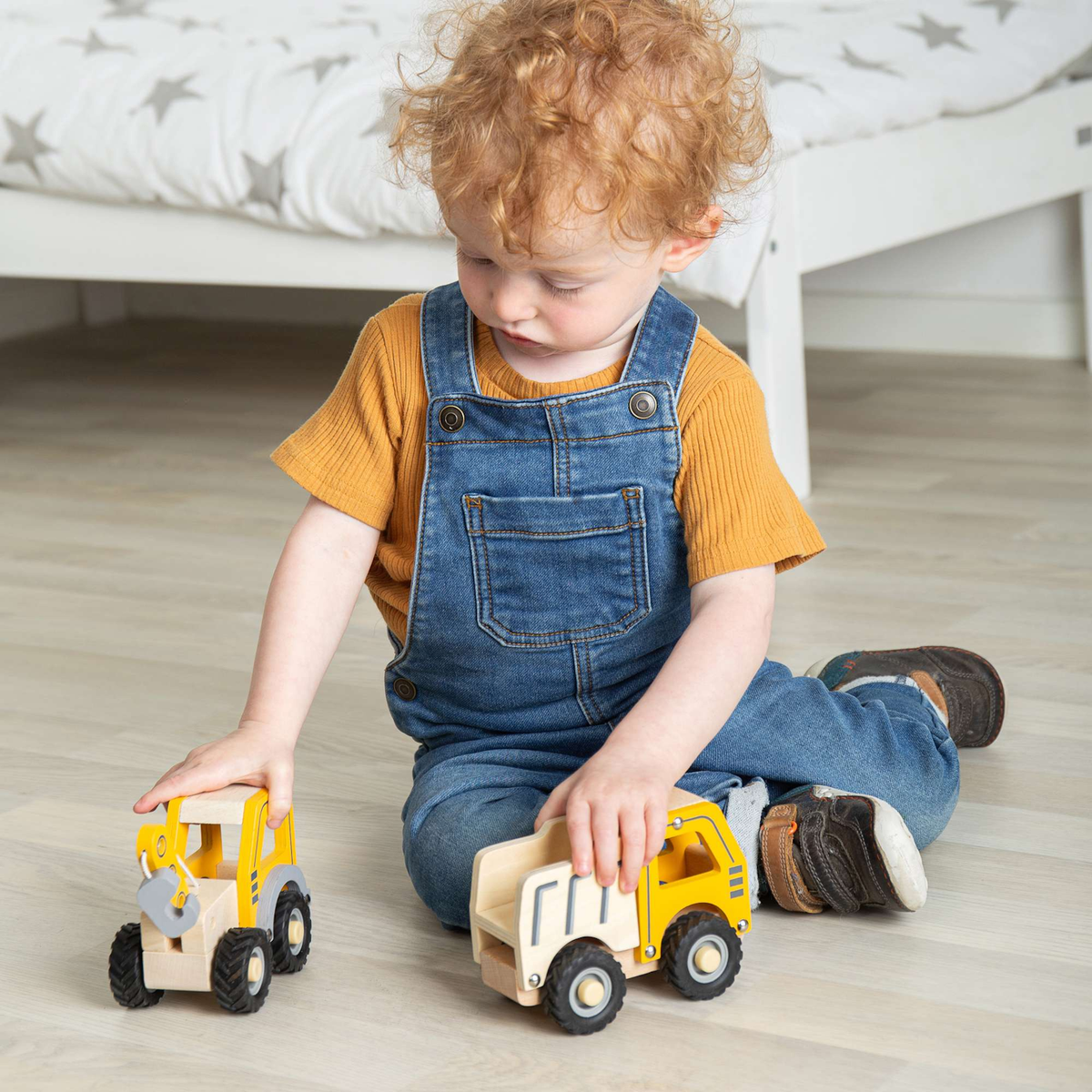 A child with curly hair, wearing blue overalls and a yellow shirt, sits on a wooden floor playing with two yellow toy trucks, focused and content.