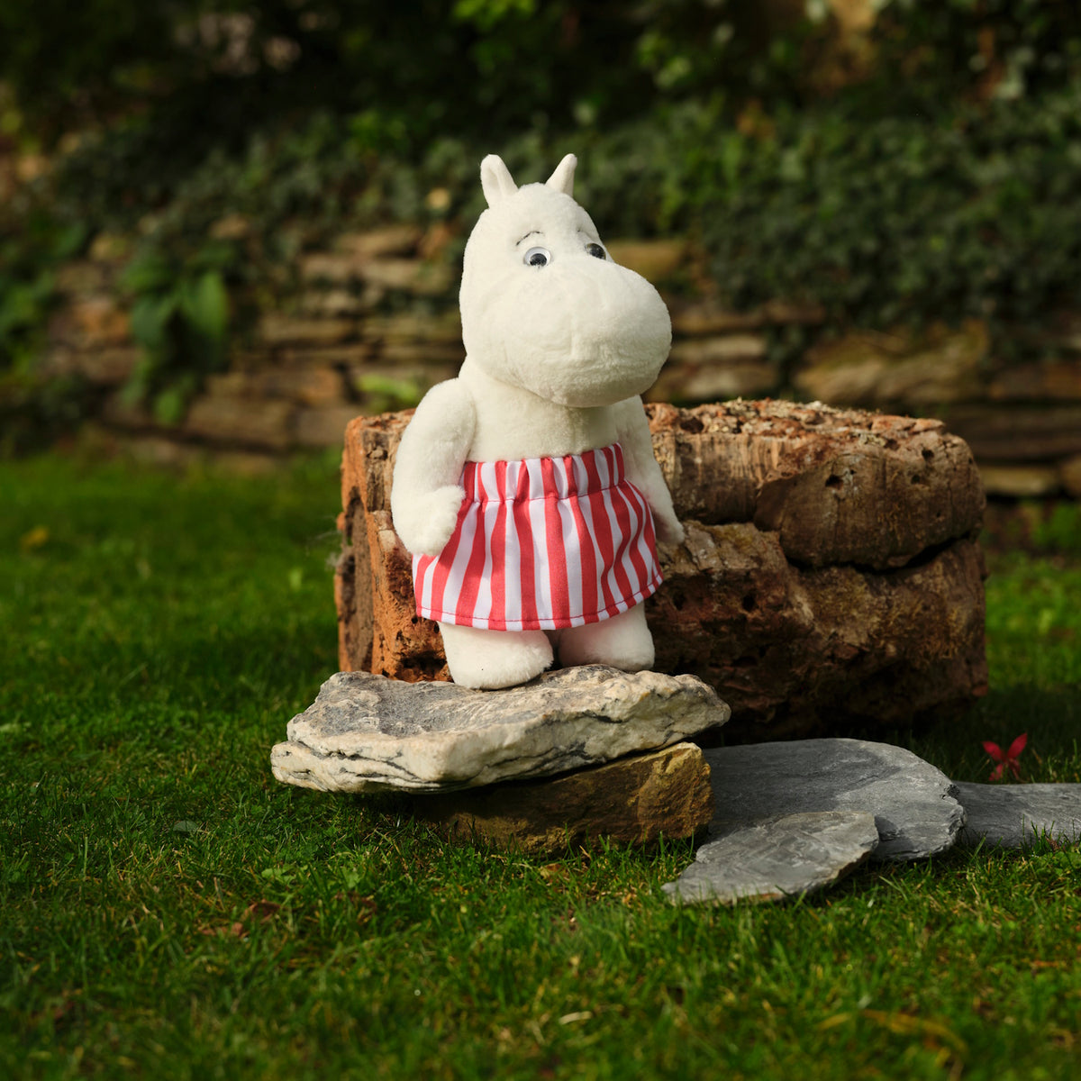 A plush toy in a red and white striped skirt stands on stones in a garden, with lush green grass and a rustic stone wall in the background.